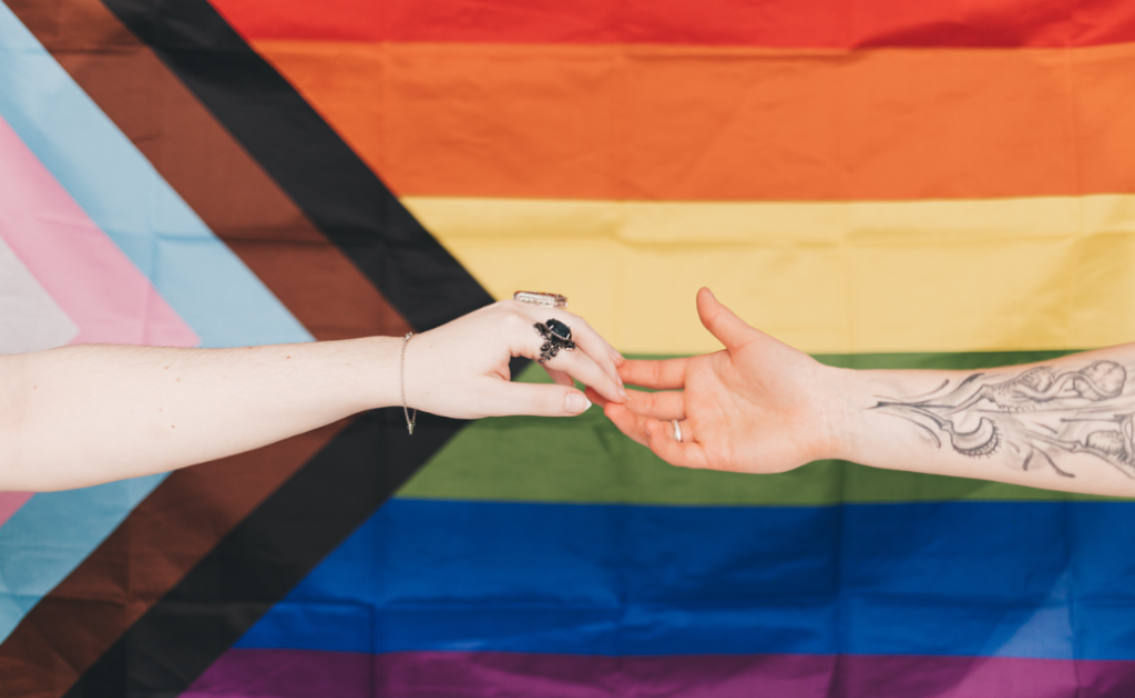 Image shows two peoples' hands reaching out towards each other with fingers touching in front of a Pride flag